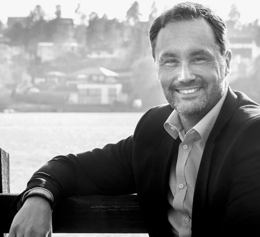 Man i kostym lutar sig mot ett räcke och ler med en sjö i bakgrunden.